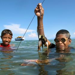 Fischen vor der Küste der Insel Atauro, Timor Leste. Zwei Jungen mit Schwimmbrillen schwimmen im türkisfarbenen Wasser und halten gefangene Fische in die Luft