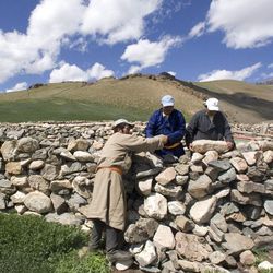 Drei Männer bauen in einer offenen, baumlosen Landschaft an einer schulterhohen Steinmauer