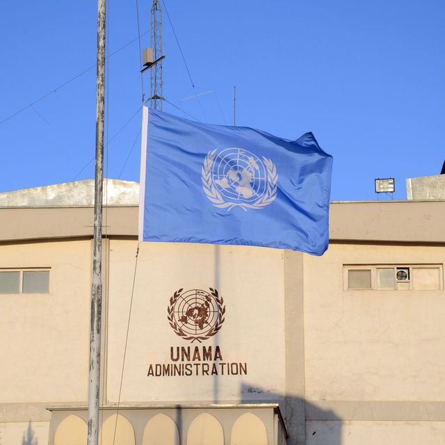 Die Flagge der Vereinten Nationen auf Halbmast am Hauptquartier der UN-Unterstützungsmission in Afghanistan (UNAMA) in Kabul. Vor einem sandfarbenen Gebäude mit dem UNAMA-Logo weht die blaue UN-Flagee auf Halbmast.