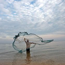 Fischer vor der Küste von Timor-Leste Bis zu den Knien steht ein Mann im flachen Küstenwasser und wirft ein großes Netz in die Höhe.