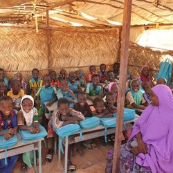 Eine Klasse von Schulkindern sitzt an Tischen in einem improvisierten Zelt aus Stroh und zeigt seine blauen Unicef-Rucksäcke.