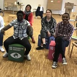 Teilnehmende am Projekt „KlimaGesichter“ im Klimahaus Bremerhaven 8 Grad Ost. Sieben international aussehende Menschen sitzen auf Würfeln, auf denen die SDGs abgebildet sind.