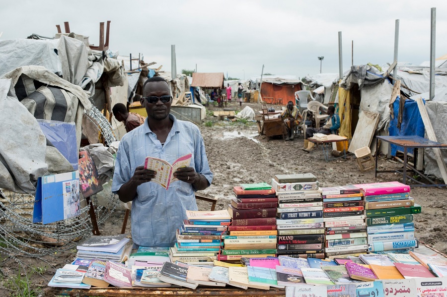 Ein afrikanischer Mann steht hinter einem Tisch voller Bücher. Er befindet sich in einer Siedlung aus improvisierten Zelten. Der Boden ist schlammig.